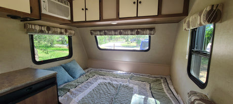 Inside a comfortable RV rental: A compact bedroom with a green quilt, blue pillows, and windows showing outdoor scenery.