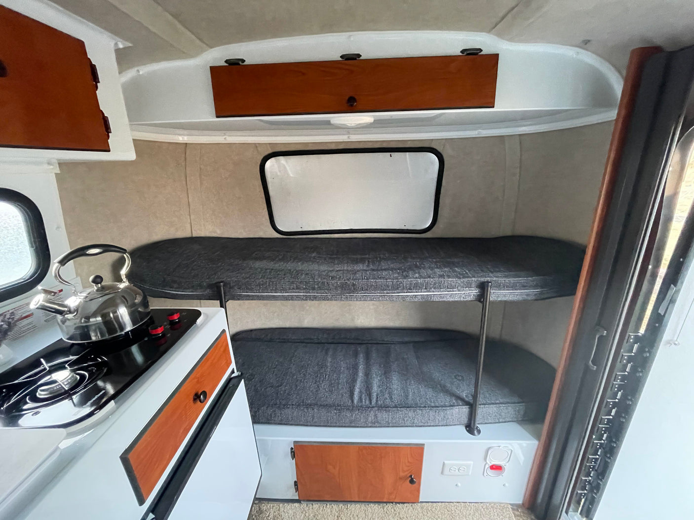 This RV rental's compact interior features bunk beds with dark cushions, a small kitchen with a stove and kettle.