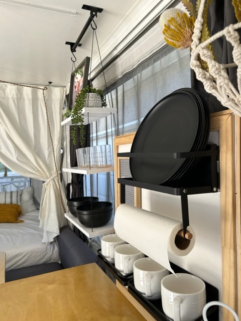 Compact RV rental interior featuring a neat kitchenette with hanging mugs, plate rack, and paper towels, next to a cozy bed. — photo 14