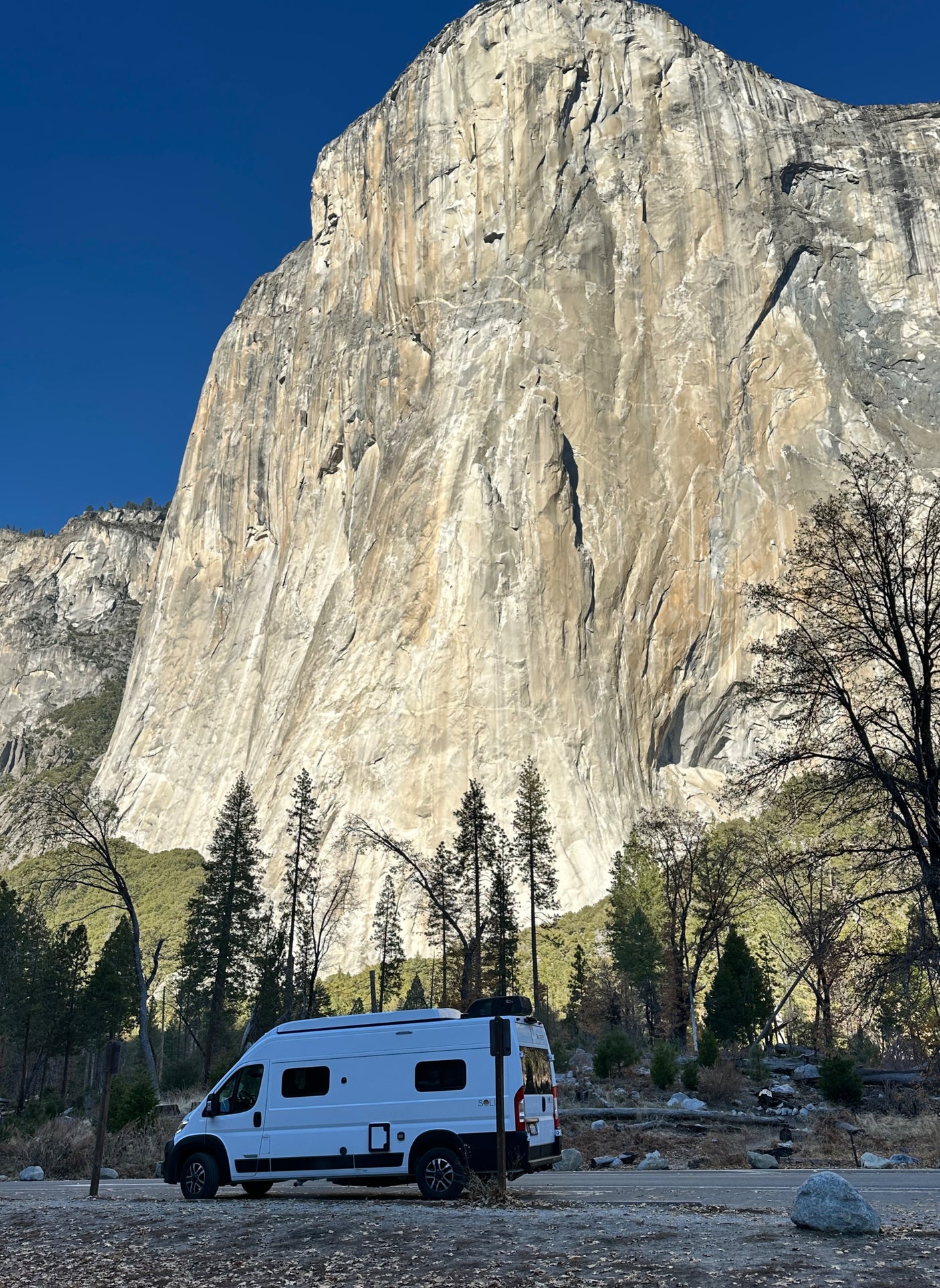 An RV rental is parked at the base of the massive El Capitan in Yosemite National Park, under a clear blue sky.