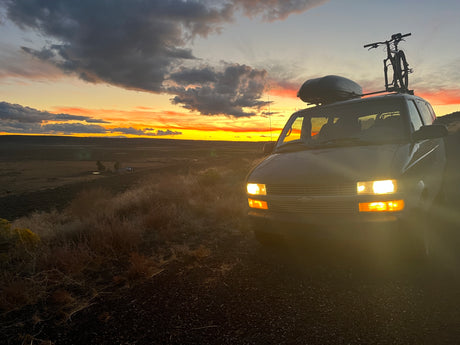 An RV rental van with a bike and cargo box sits under a vibrant sunset over a wide, arid landscape.