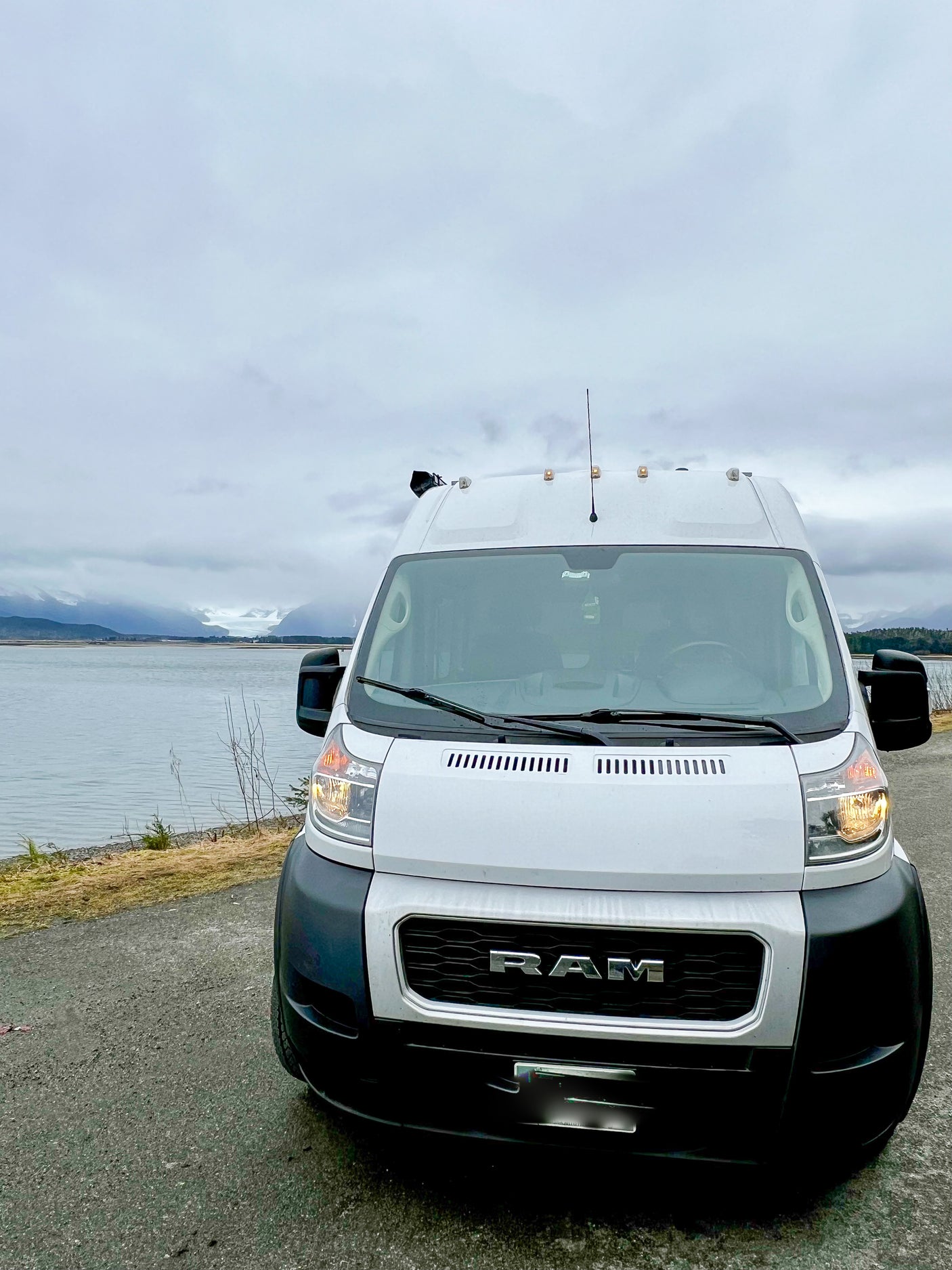 White Ram ProMaster RV rental by a scenic lake with distant snow-capped mountains & glacier under cloudy skies.