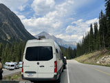 An rv rental van with Indiana plates rests by a mountain road, with other RVs parked nearby in a stunning landscape.