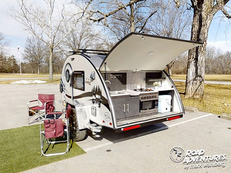 Teardrop travel trailer RV rental parked outdoors, rear hatch open to reveal an outdoor kitchen, stove, microwave, and chairs.