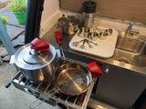 Compact RV rental kitchen with pull-out stovetop, pots, pan, measuring tools, Stanley mug & sink for outdoor cooking.