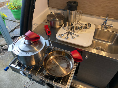 Compact RV rental kitchen with pull-out stovetop, pots, pan, measuring tools, Stanley mug & sink for outdoor cooking.