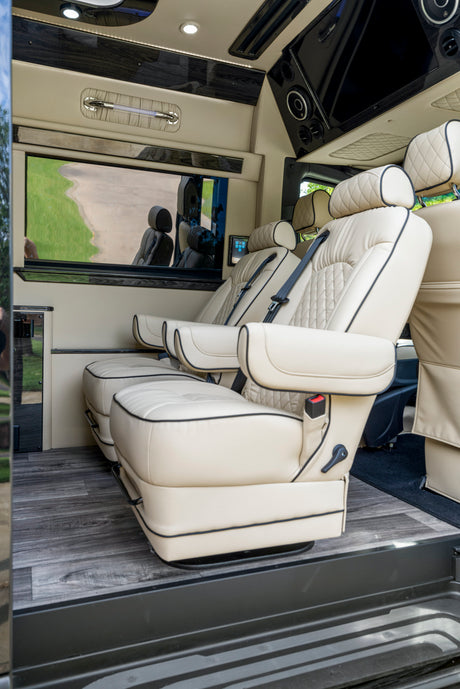 Plush interior of a custom RV rental, showing cream leather captain's chairs, wood floors, and entertainment system.
