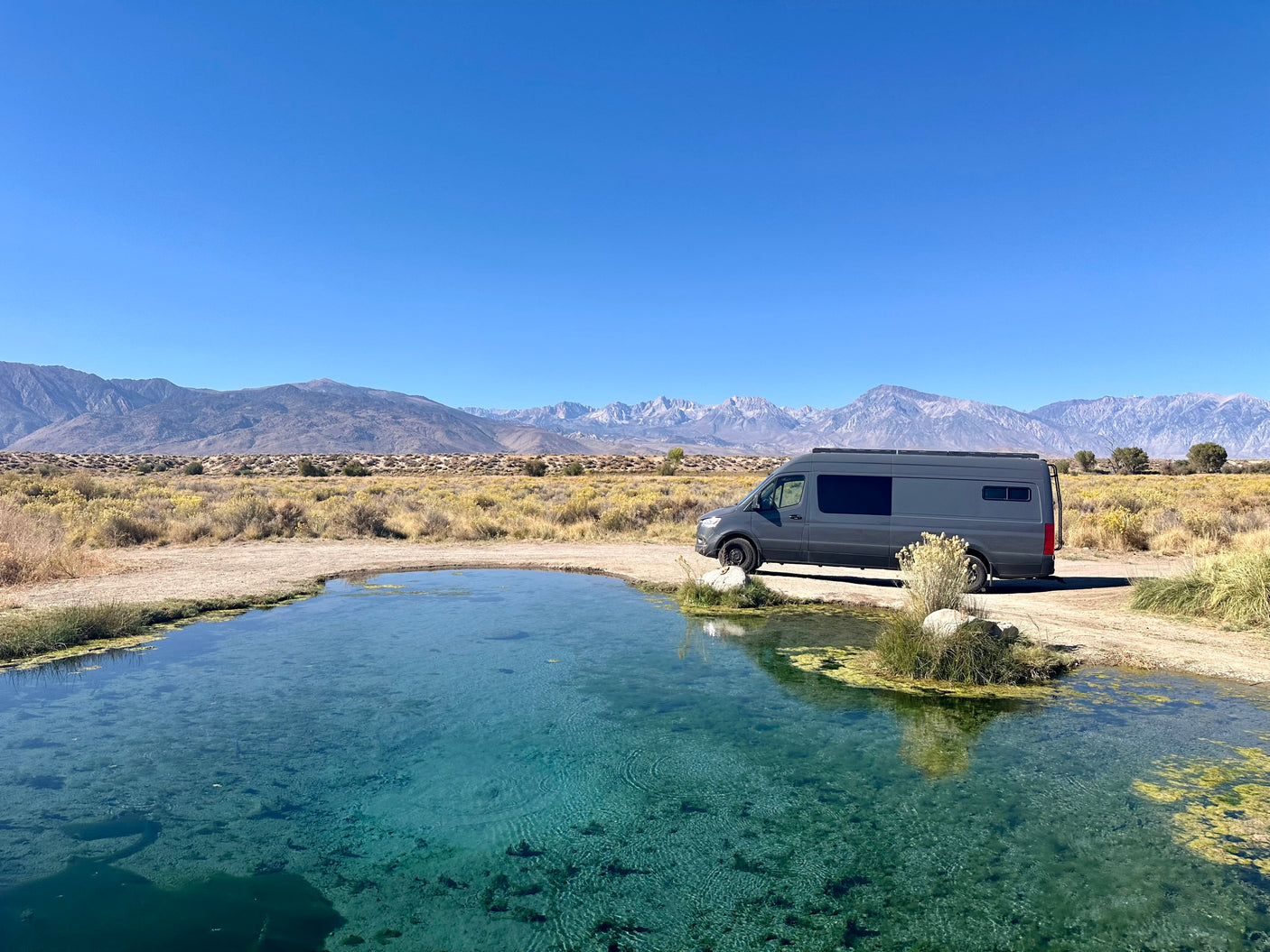 Grey RV rental next to a crystal-clear blue pond, with expansive desert landscape and majestic mountains in the background.