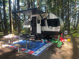 RV rental travel trailer campsite with extended awning, picnic area, grill, & dog under tall forest trees.