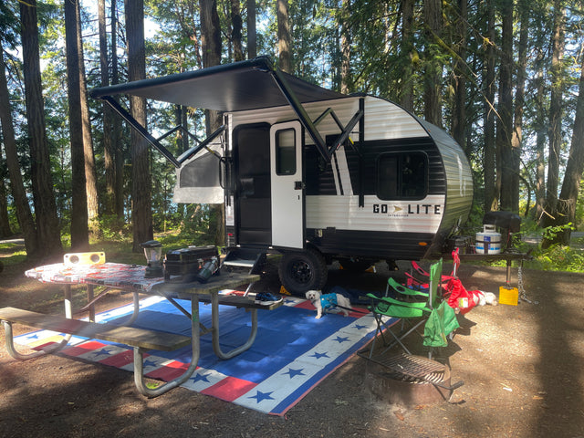 RV rental travel trailer campsite with extended awning, picnic area, grill, & dog under tall forest trees.