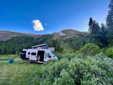 RV rental camper van with pop-top raised, nestled in a mountain forest clearing under a blue sky.