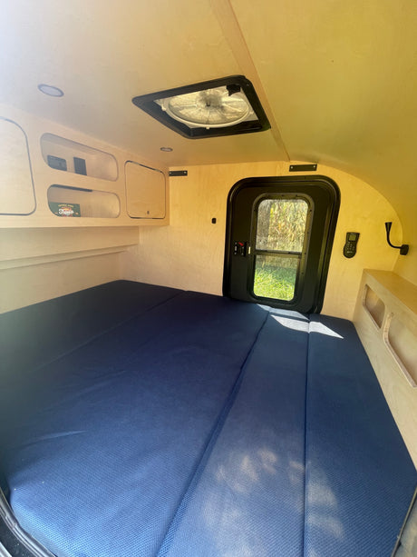 A cozy RV rental bedroom interior with a large blue mattress, built-in wooden storage, and a ceiling fan.