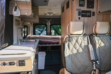 This RV rental's interior features a compact kitchen with sink, cozy bed, & controls near passenger seats.