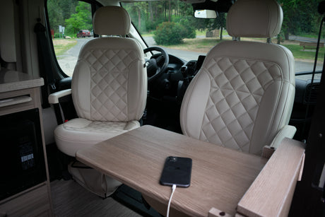 Inside a modern RV rental, featuring luxurious quilted seats, a fold-out table with a charging phone, and a view of the road ahead.