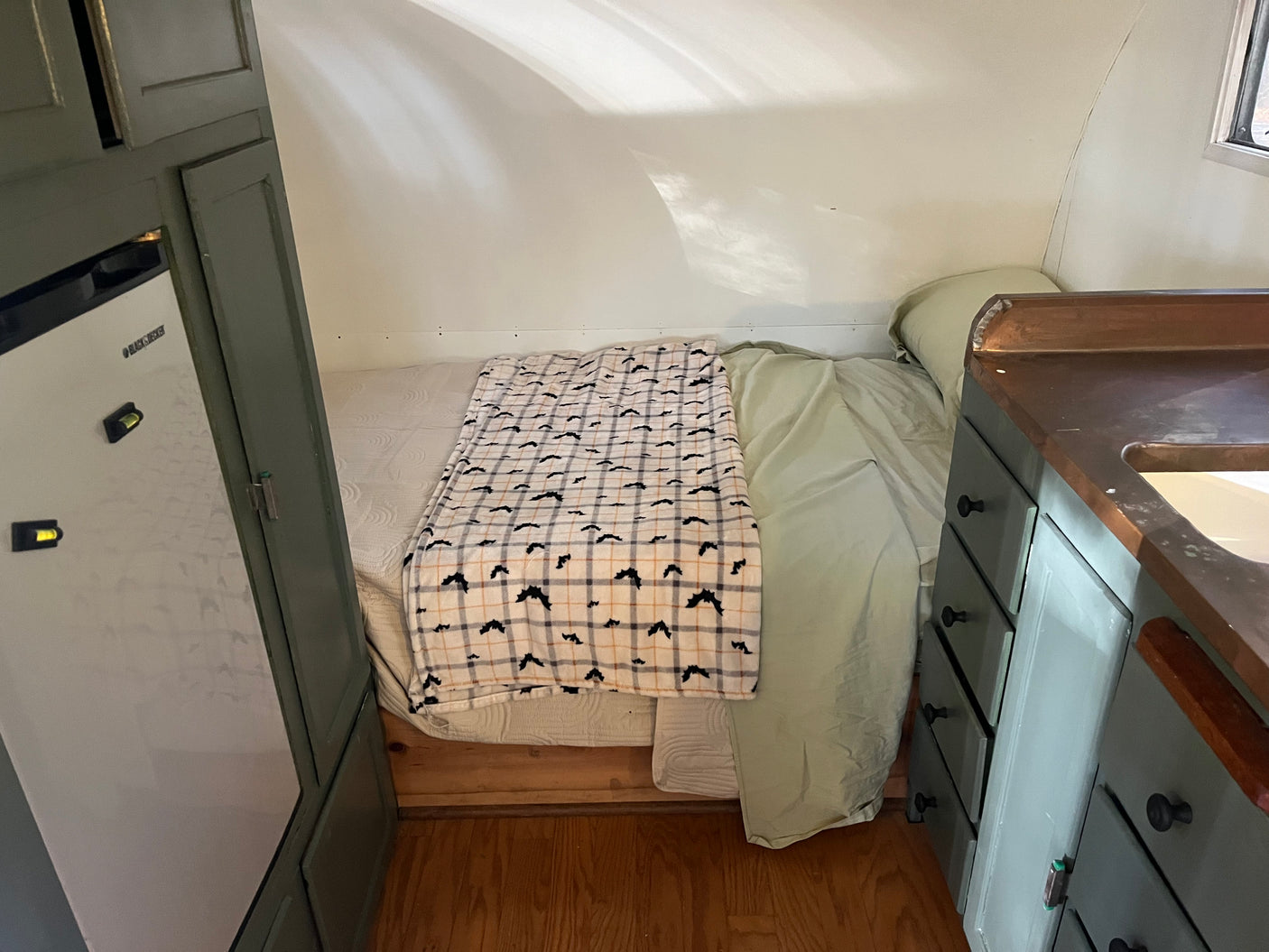 Cozy RV rental interior with a bed, bat-patterned blanket, compact fridge, and a small kitchenette with wooden counters.