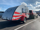 A silver T@G teardrop RV with red stripes, ideal for an rv rental, is hitched to an SUV in a sunny parking lot.