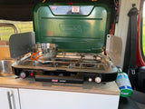 RV rental: Coleman propane stove with pot set up for cooking inside the camper. A green propane tank is nearby.