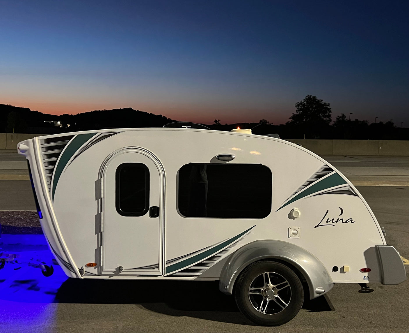 White Luna tear-drop RV rental for tiny camping, accented with teal stripes and blue underglow at dusk.