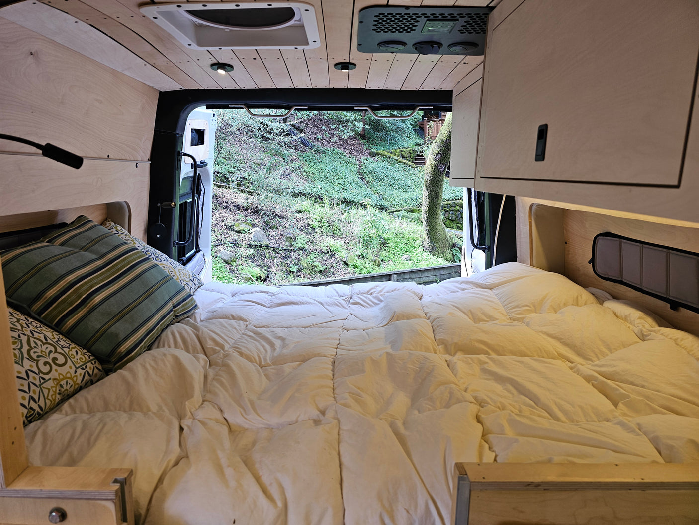 Relax in this cozy RV rental! Enjoy the comfy bed and stunning view of a lush green forest through the open back doors.