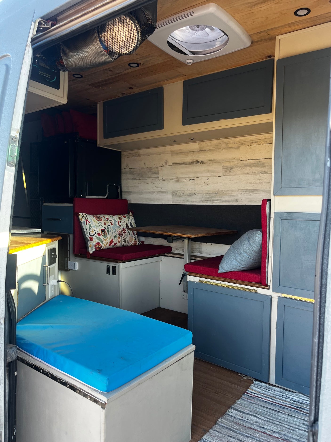 View inside an RV rental, featuring a dinette with red cushions, an adjustable table, overhead storage, and a blue bed.