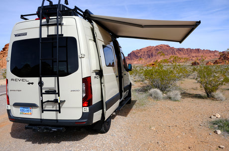 Winnebago Revel rv rental van parked in a desert landscape with red rock formations, its awning extended.