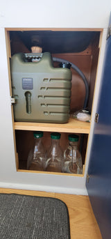 RV rental: Under-sink view showing a large green water tank (UPWOIGH) & glass bottles for compact, off-grid water storage.