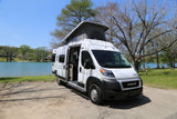 White pop-top RV rental parked lakeside, door open to serene blue water and green trees. Perfect for adventure.