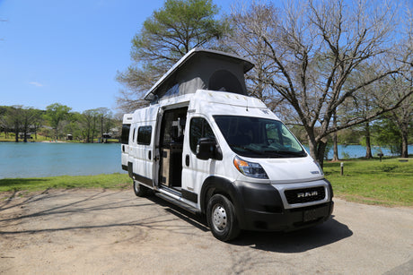 White pop-top RV rental parked lakeside, door open to serene blue water and green trees. Perfect for adventure.