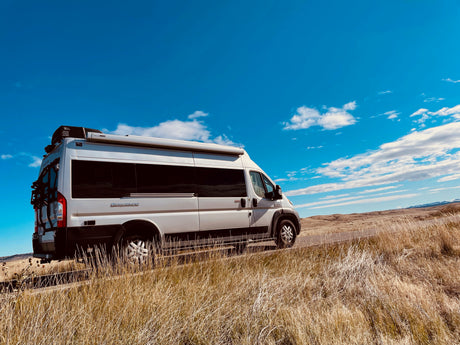Silver rv rental van on a golden grass roadside. Blue sky with clouds, distant hills.