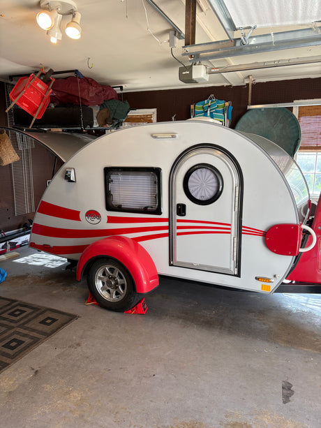 A sleek white T@G teardrop trailer with red stripes, perfect for an RV rental, parked inside a garage.