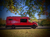 Bright red RV rental van parked on asphalt next to grass, framed by autumn leaves & blue sky.