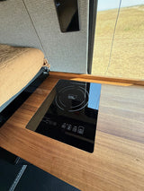 Inside an RV rental: a modern induction cooktop built into a wood counter, next to a comfy bed with a view of nature.