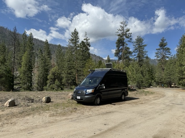 Dark blue rv rental van with solar panels parked in a forest of tall pine trees under a blue sky with white clouds.|RV rental: panoramic views of hot springs & snowy mountains from inside a camper van's kitchen while cooking.|Dark blue Ford Transit camper van (RV rental) parked on a dirt path with a clear ocean view under a blue sky.|Inside this RV rental: a camper van interior with wood flooring — photo 1
