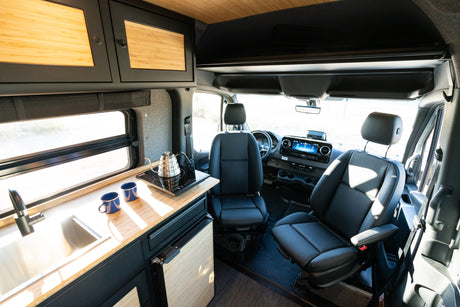 Modern RV rental interior: kitchen with sink, cooktop, and comfy swivel seats upfront. Ready for your next adventure.
