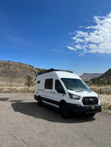 White Ford Transit campervan RV rental with roof awning, parked on asphalt in a scenic mountainous landscape under a blue sky.