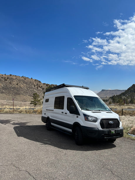 White Ford Transit campervan RV rental with roof awning, parked on asphalt in a scenic mountainous landscape under a blue sky.