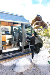 Experience van life in an RV rental. Woman by an open camper van with an awning, kitchen, and seats in a desert landscape.