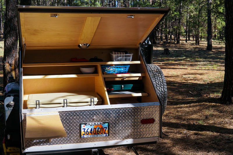 RV rental teardrop trailer's open galley, featuring an outdoor kitchen with organized camping gear in a pine forest. — photo 5