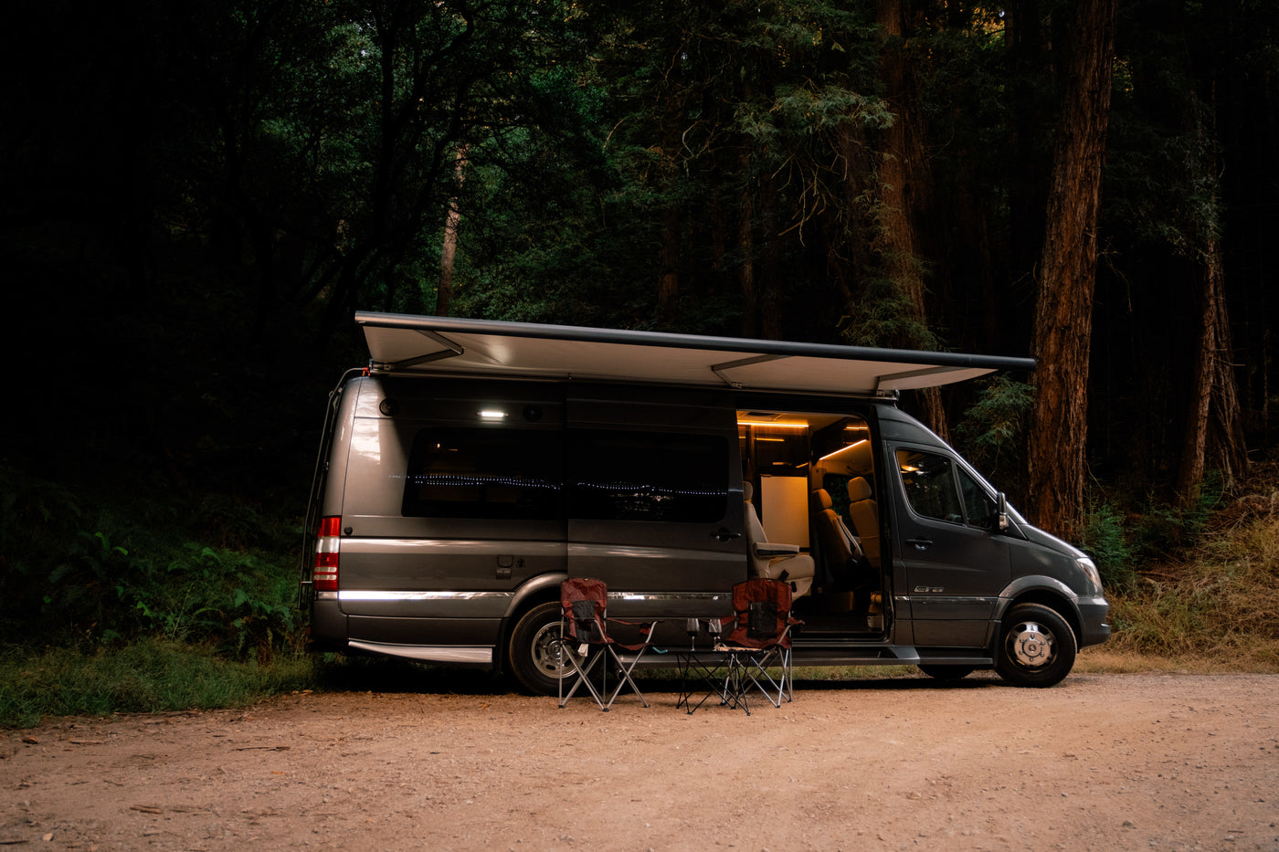 Cozy dark gray RV rental campervan with illuminated interior, extended awning, and chairs outside on a forest dirt road at dusk. — photo 17