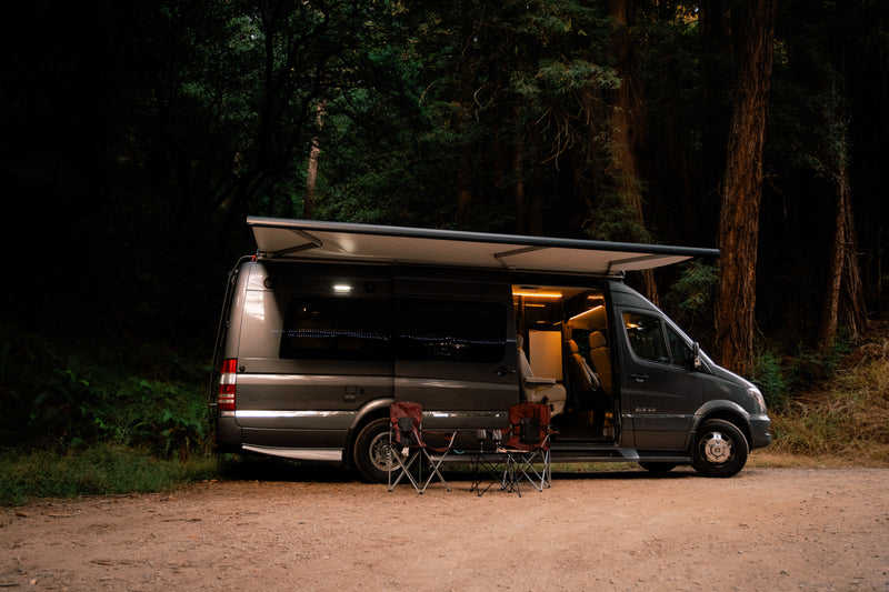 Cozy dark gray RV rental campervan with illuminated interior, extended awning, and chairs outside on a forest dirt road at dusk. — photo 17
