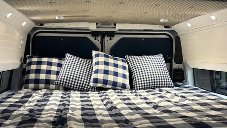Inviting RV rental bed with blue & white checkered bedding/pillows, illuminated by recessed lights in a camper van.