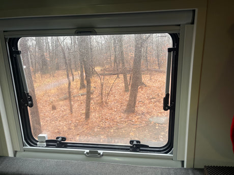 View from an RV rental window on a rainy day. A forest with bare trees and autumn leaves is seen through the wet glass.