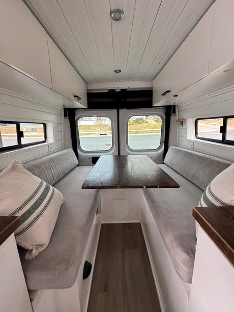 A bright, white RV rental interior with shiplap walls, a wooden dining table, comfy gray benches, and storage, perfect for travel.