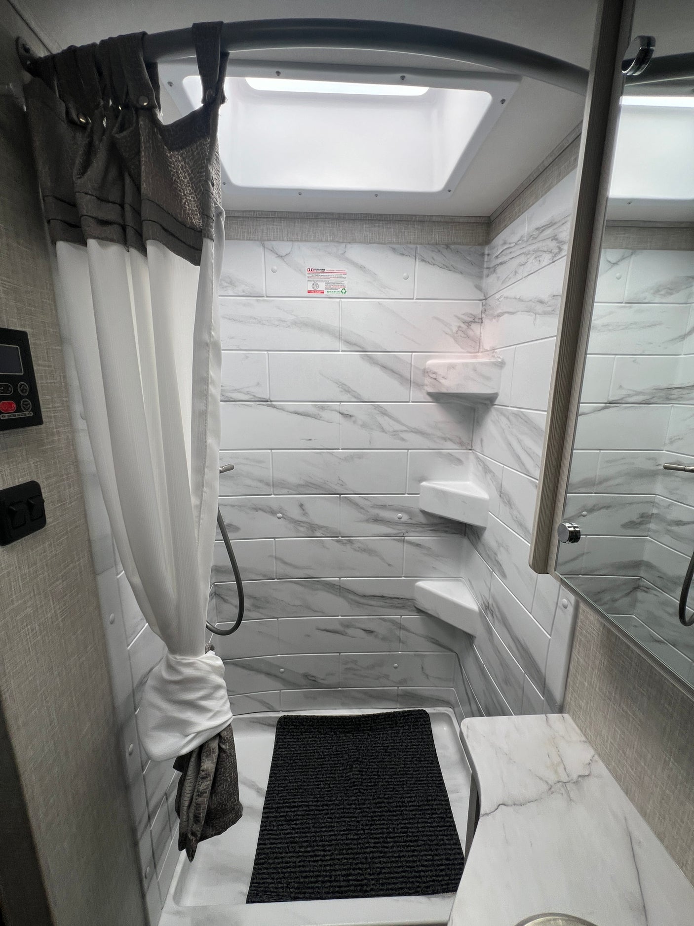 Bright, compact RV rental shower with white tiled walls, corner shelves, a skylight, and a sleek gray and white curtain.