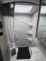Bright, compact RV rental shower with white tiled walls, corner shelves, a skylight, and a sleek gray and white curtain.