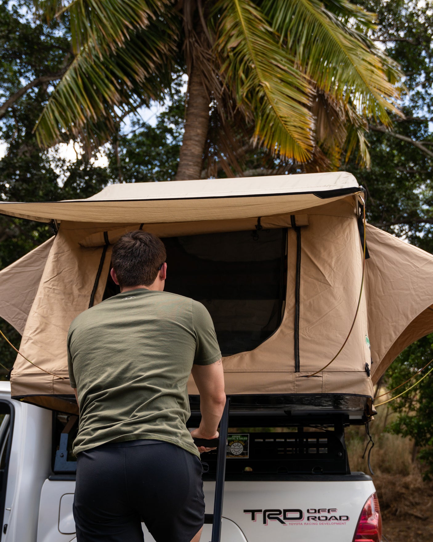 A man climbs into a beige rooftop tent on a white TRD pickup truck, part of an RV rental setup for camping.