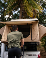 A man climbs into a beige rooftop tent on a white TRD pickup truck, part of an RV rental setup for camping.