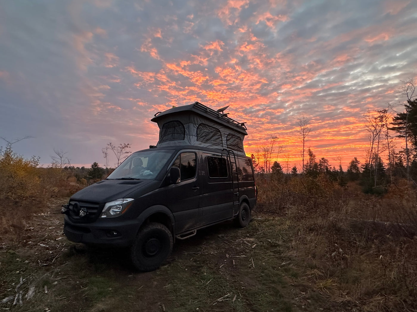 A dark pop-top camper van parked in a field, bathed in the fiery orange and pink hues of a sunset. Great for an rv rental adventure. — photo 6