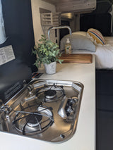 Modern RV rental: a compact kitchen setup with a stainless steel two-burner stove, sink, and a comfortable bed.