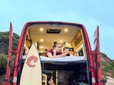 Enjoying van life in an RV rental: A woman and child lounge inside their red camper van, surfboard included. Perfect for family travel.